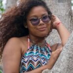 Woman with curly hair and sunglasses posing against a tree trunk outdoors.