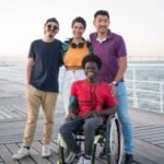 Diverse group of friends happily posing by the sea on a sunny day, with one using a wheelchair.