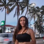 Portrait of a smiling young woman outdoors with palm trees and city backdrop at twilight.